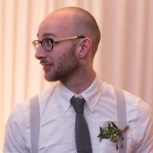A bald man wearing glasses and suspenders displays available .COM domains.